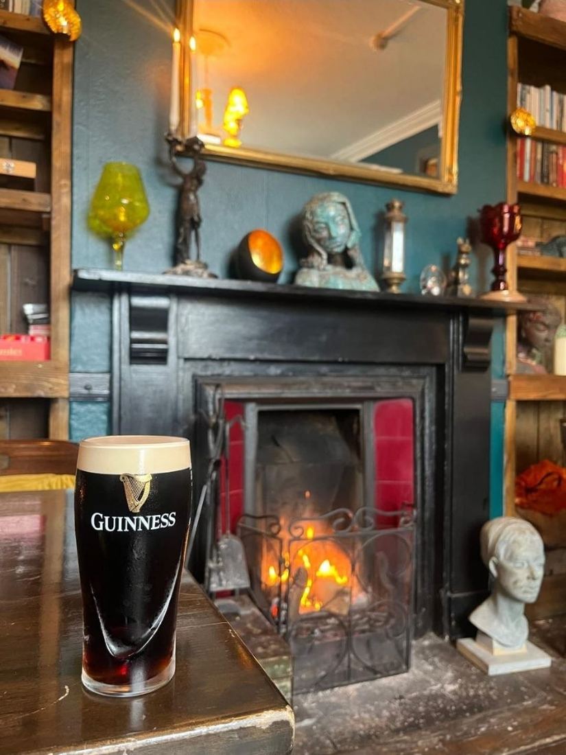 A pint of stout on a table in front of the lit fire, under the gilt mirror and the mantel of curiosities.